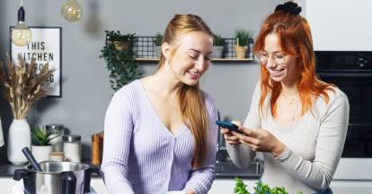 Isy und Tessa stehen hinter einer Arbeitsfläche und schauen auf ein Handy.