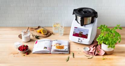 Monsieur Cuisine auf einer Arbeitsplatte, daneben ein Kochbuch, aufgeschlagen und ein Topf mit Kräutern