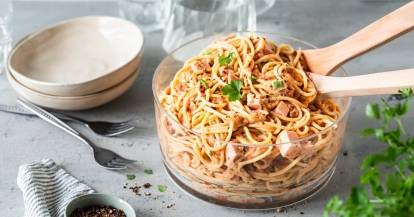 Eine Glasschale mit Spaghettisalat auf grauer Arbeitsplatte, daneben Teller und KrÀuter