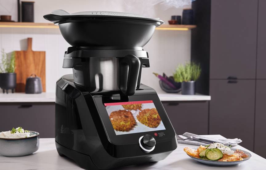 Monsieur Cuisine smart in der Black Edition auf einer Arbeitsplatte, daneben Kartoffelpuffer auf einem Teller.