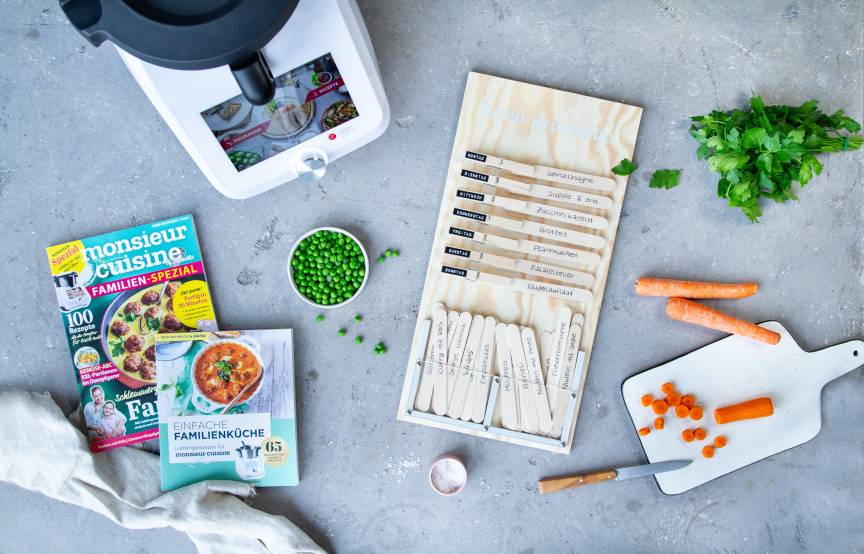 Auf einer Arbeitsplatte liegen ein Speiseplan, ein Familien-Kochbuch, das neue Familien-Spezial, der Monsieur Cuisine sowie einige frische Lebensmittel