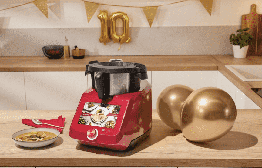 Der Monsieur Cuisine Smart in rot auf einer Holzarbeitsplatte, daneben zwei goldene Luftballons